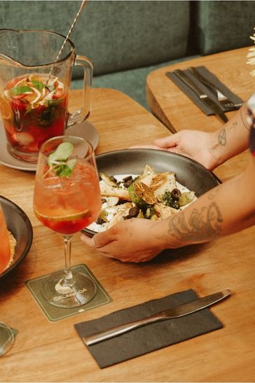 A woman stands holding a plate filled with food and drinks, showcasing her culinary preparation with a cheerful expression.