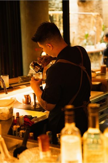 A bartender in an apron prepares a cocktail at the bar, focused on creating a perfect drink for customers