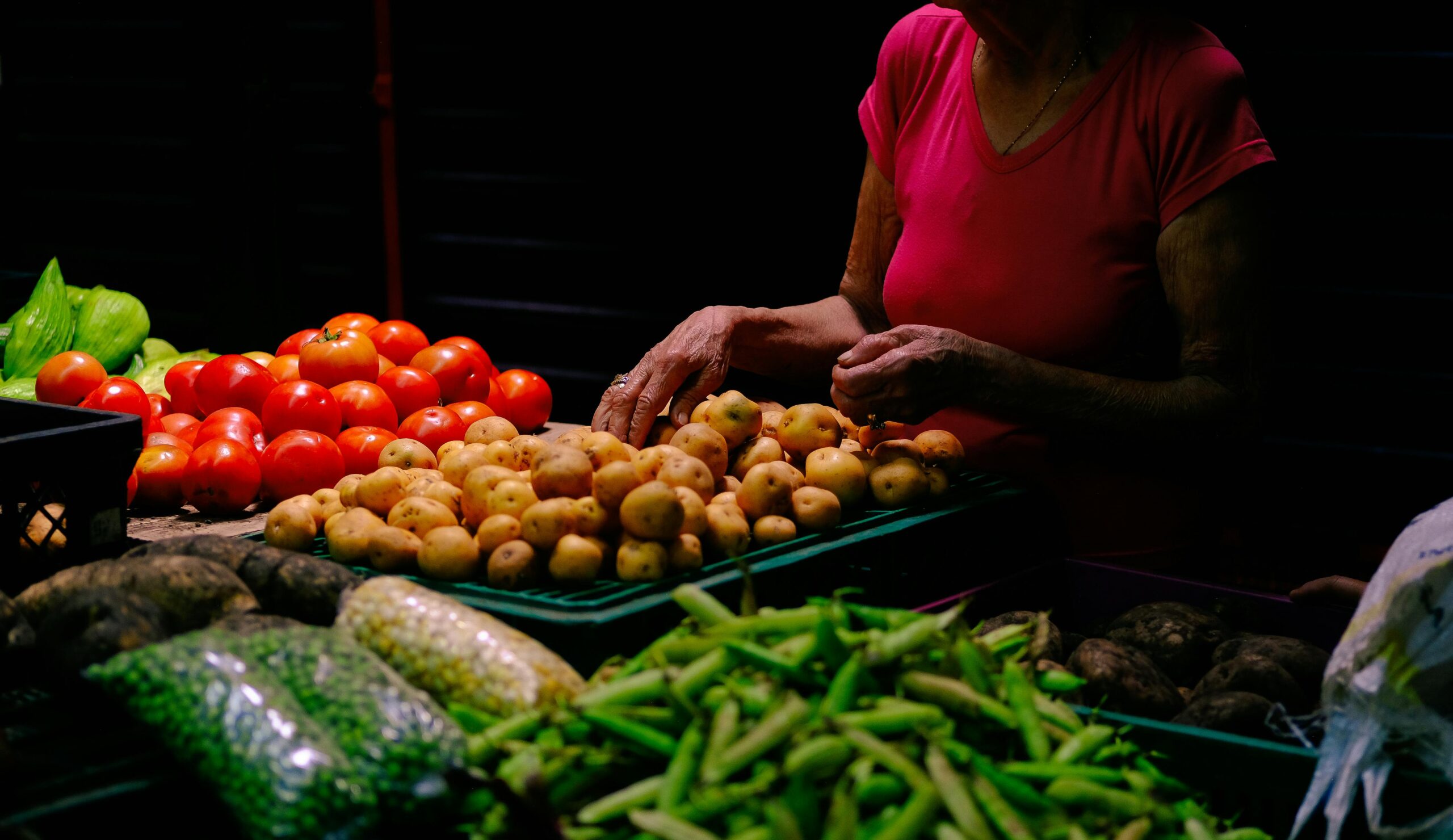A woman stands by a table filled with various fresh vegetables, smiling as she prepares to cook.