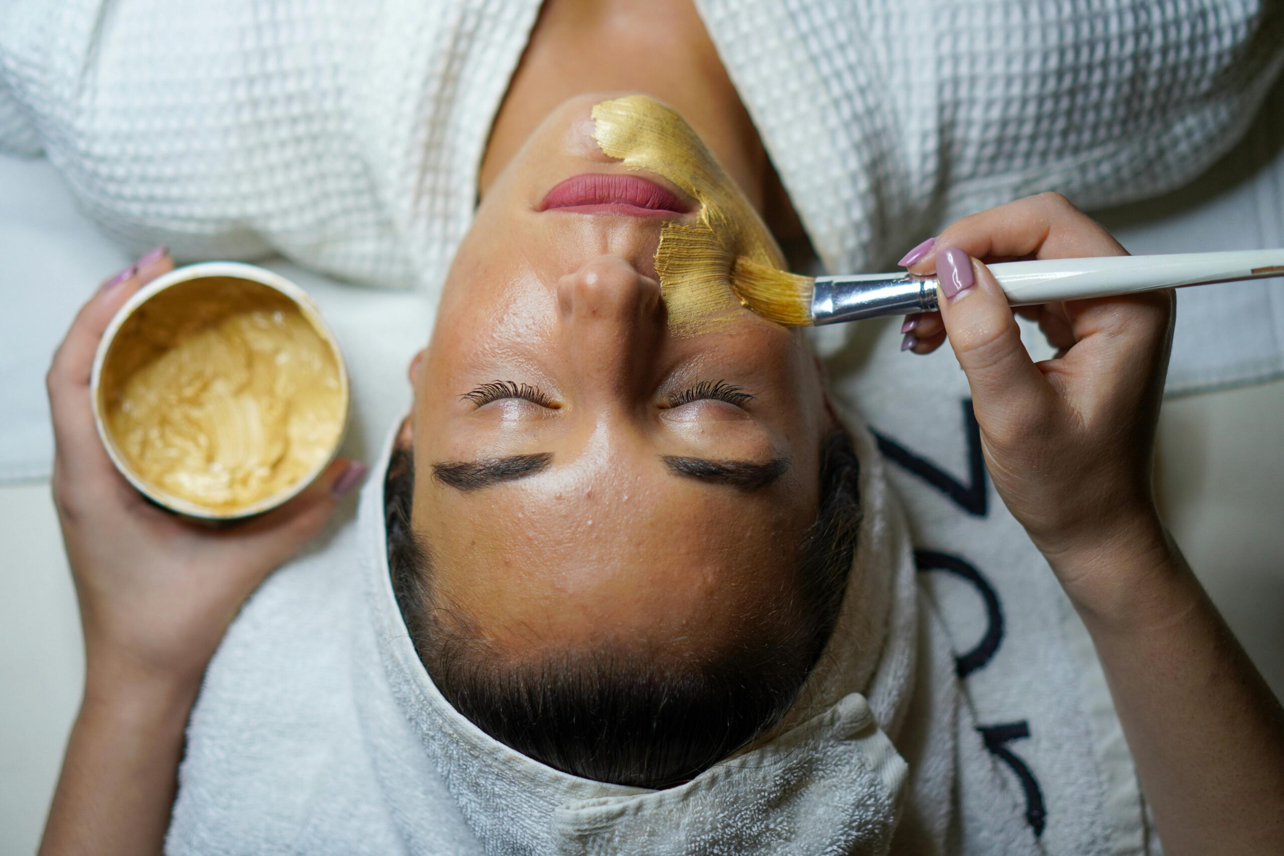 A woman receiving a facial treatment as a therapist applies product with a brush on her face.