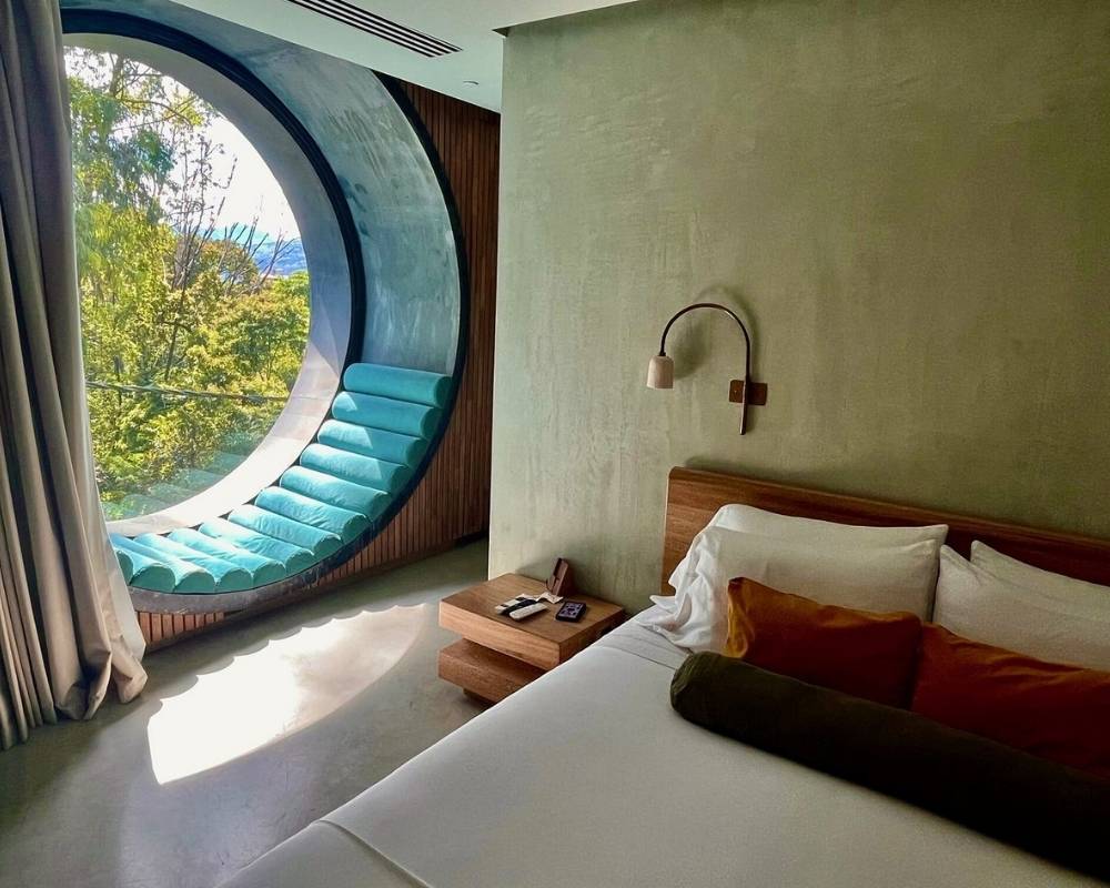 Dormitorio con una ventana circular y una cama, decorado con tonos suaves y luz natural que entra por la ventana.