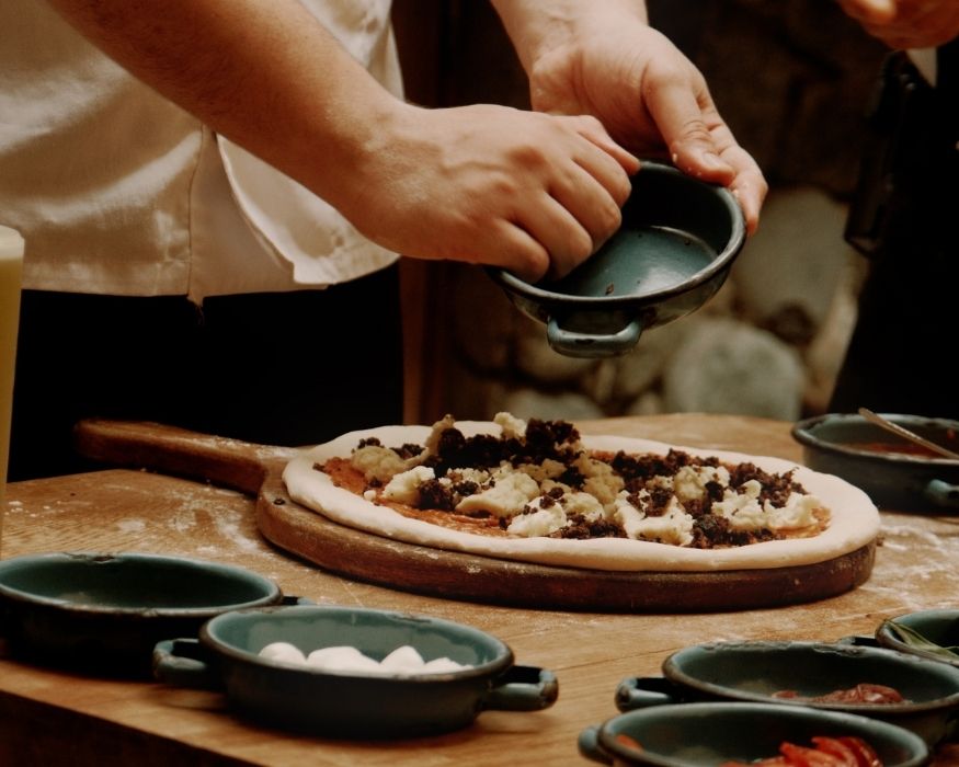 Un hombre está colocando coberturas sobre una pizza recién hecha