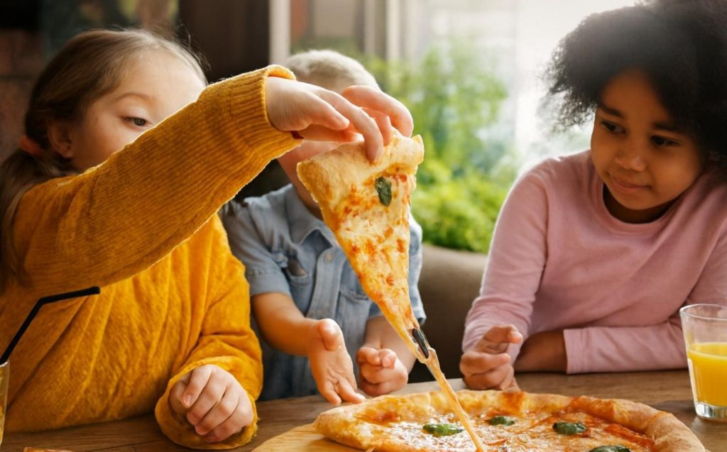 Tres niños disfrutan de pizza en una mesa, sonriendo y compartiendo un momento divertido en un restaurante familiar.