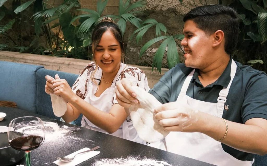 Two people sitting at a table, preparing pizza dough together with smiles on their faces.