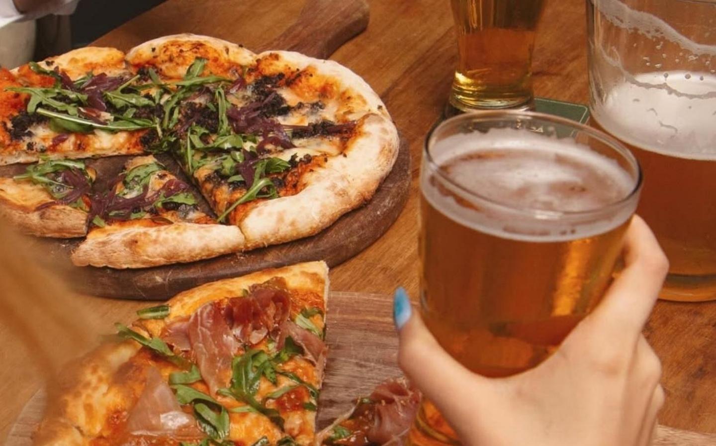 A person enjoying a slice of pizza while holding a glass of beer, smiling in a casual setting